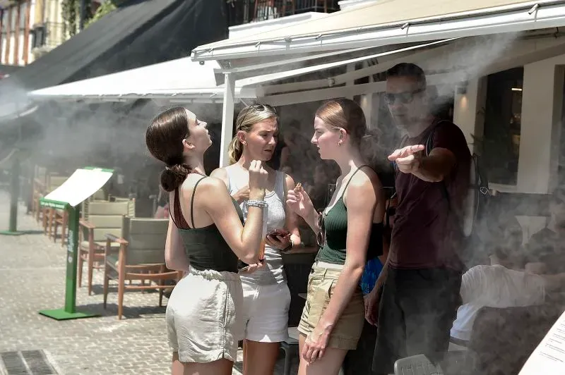 مجموعة من النساء يتحدثن في حديقة عامة بأثينا، بينما يتصاعد بخار الماء من نظام تبريد لمواجهة الحرارة الشديدة.