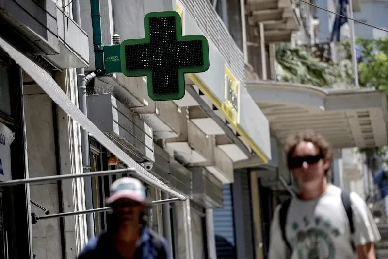 شاشة عرض درجة حرارة في أثينا تسجل 44 درجة مئوية، مع مشاة يتجولون في الشارع. تعكس الصورة تأثير موجات الحر على المدينة.