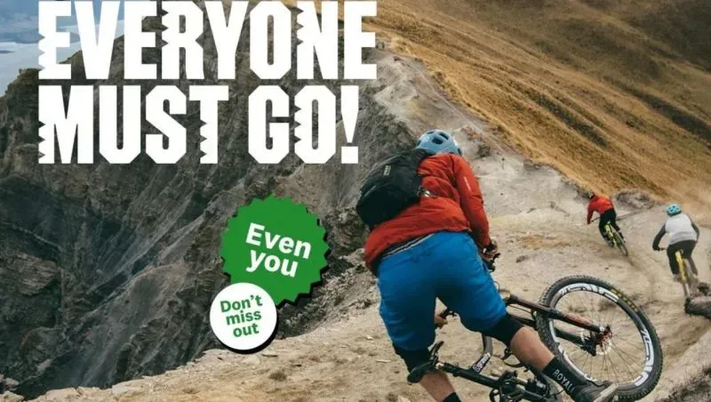 ملصق حملة السياحة النيوزيلندية يظهر راكبي دراجات جبلية في مناظر طبيعية خلابة، مع شعار "يجب أن يذهب الجميع!" لجذب السياح.