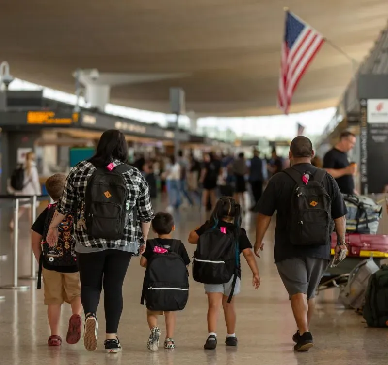 عائلة مكونة من خمسة أفراد تسير في مطار مزدحم، تحمل حقائب ظهر، متجهة نحو مغادرتها الولايات المتحدة إلى المكسيك.