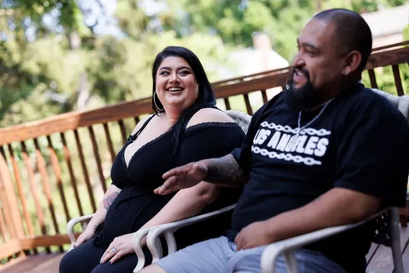 ساشا وخوليو يجلسان معًا على شرفة، يضحكان ويتبادلان الأحاديث، مع خلفية خضراء. يعكسان تحديات الحياة كزوجين من خلفيات مختلفة.