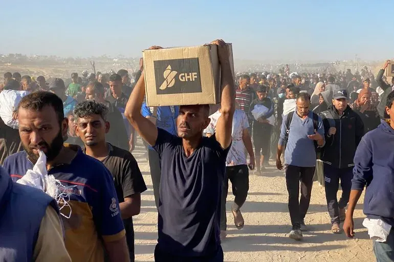 مواطن يحمل صندوق مساعدات من مؤسسة GHF وسط حشود من الأشخاص في غزة، يعكس مشهد التوتر والبحث عن المساعدة الإنسانية.