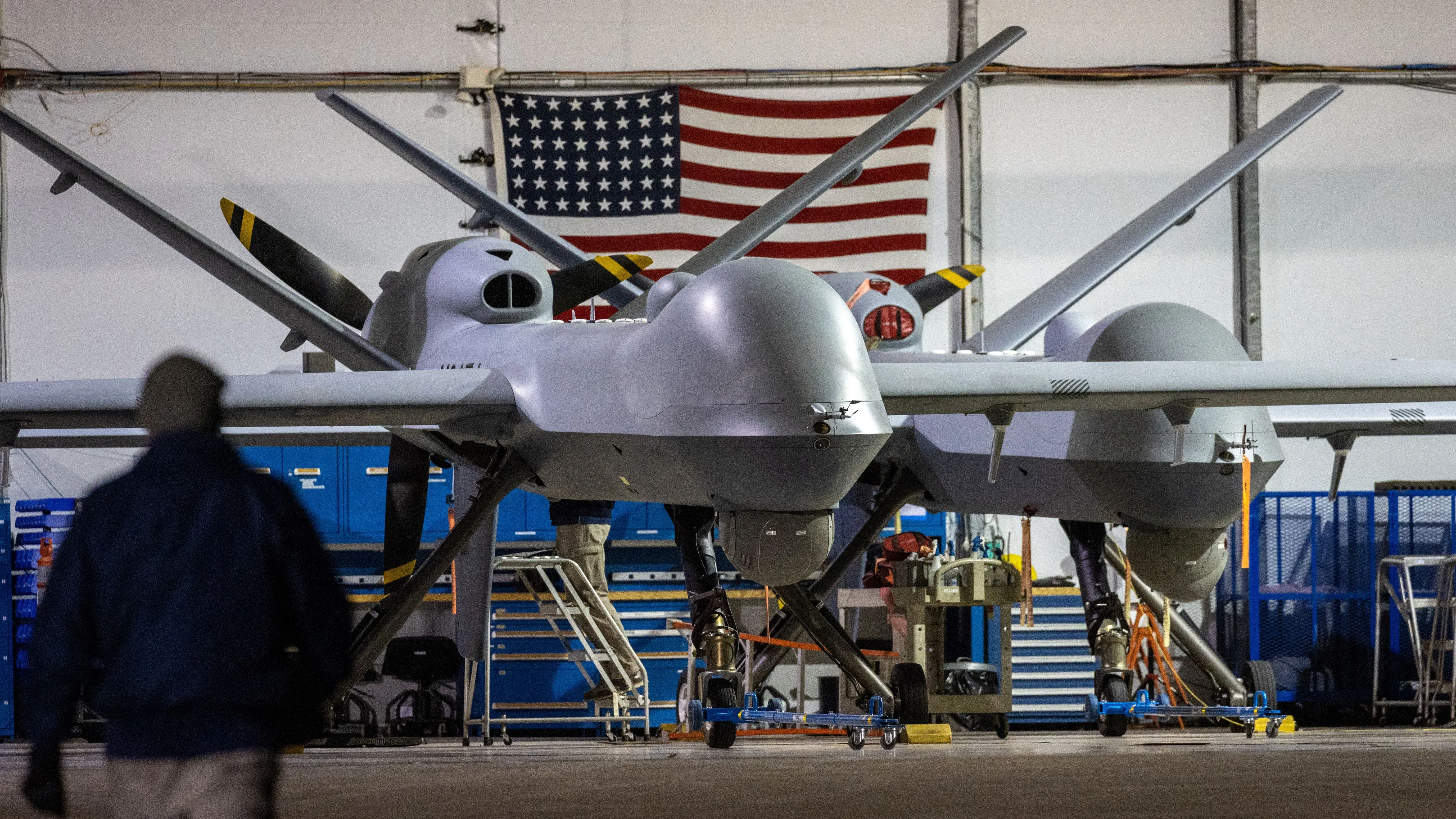 طائرات MQ-9 ريبر بدون طيار داخل قاعدة عسكرية أمريكية، مع العلم الأمريكي في الخلفية، تشير إلى أنشطة التجسس على عصابات المخدرات.