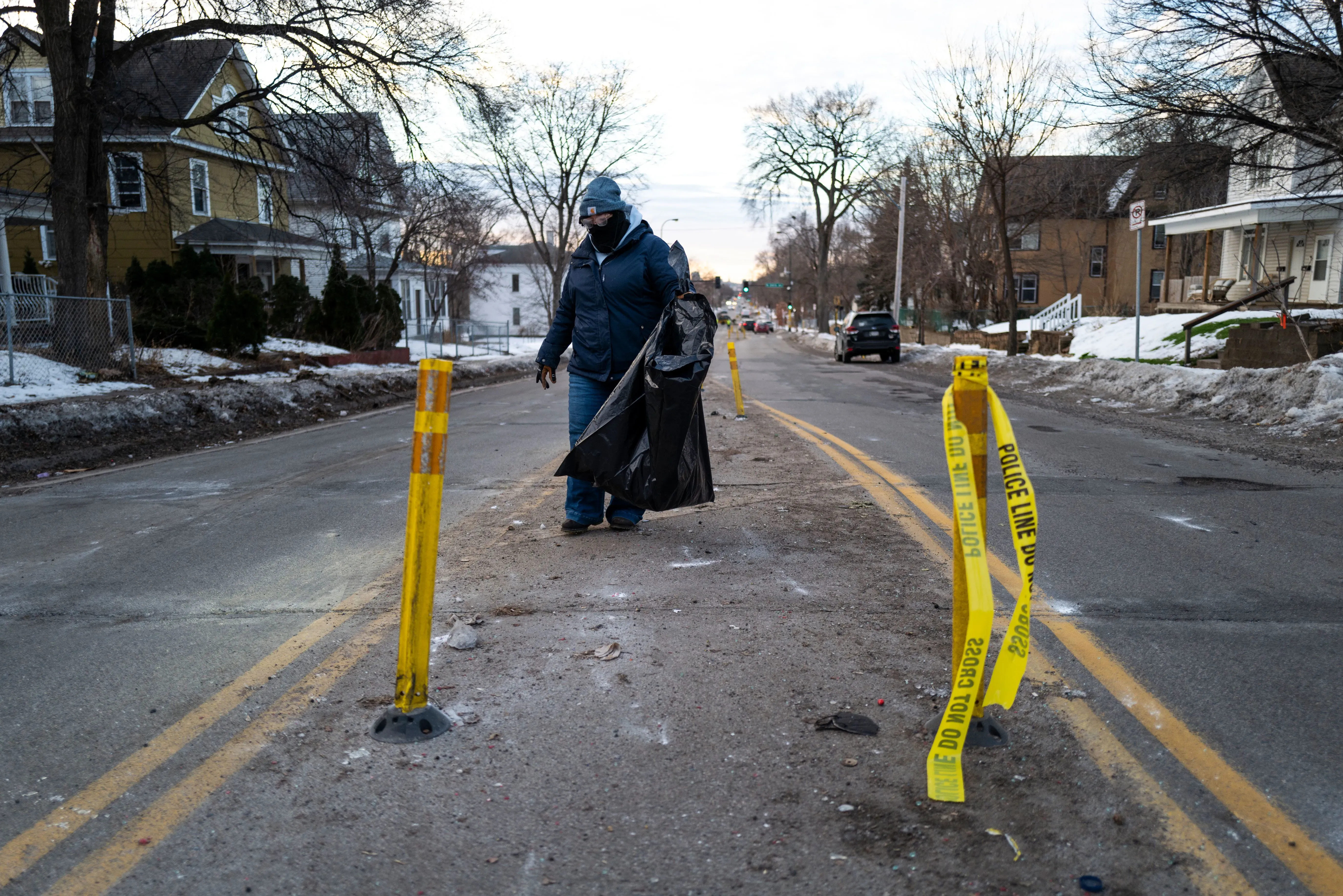 شخص يجمع القمامة في شارع مغلق بشريط شرطة أصفر، مع بقايا ثلوج وأشجار في الخلفية، مما يعكس جهود التنظيف في منطقة متأثرة بالاضطرابات.