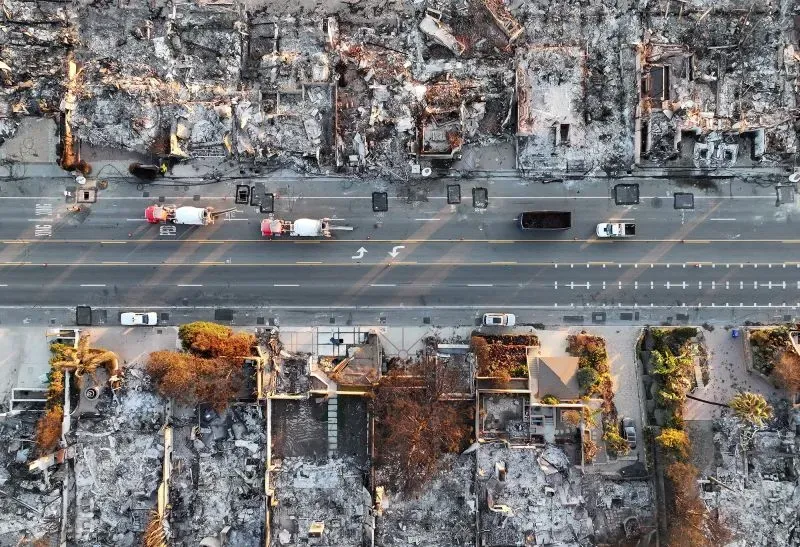مشهد جوي لشارع في لوس أنجلوس، حيث تظهر المنازل المحترقة والمدمرة جراء حرائق الغابات، مع وجود شاحنات إطفاء على الطريق.