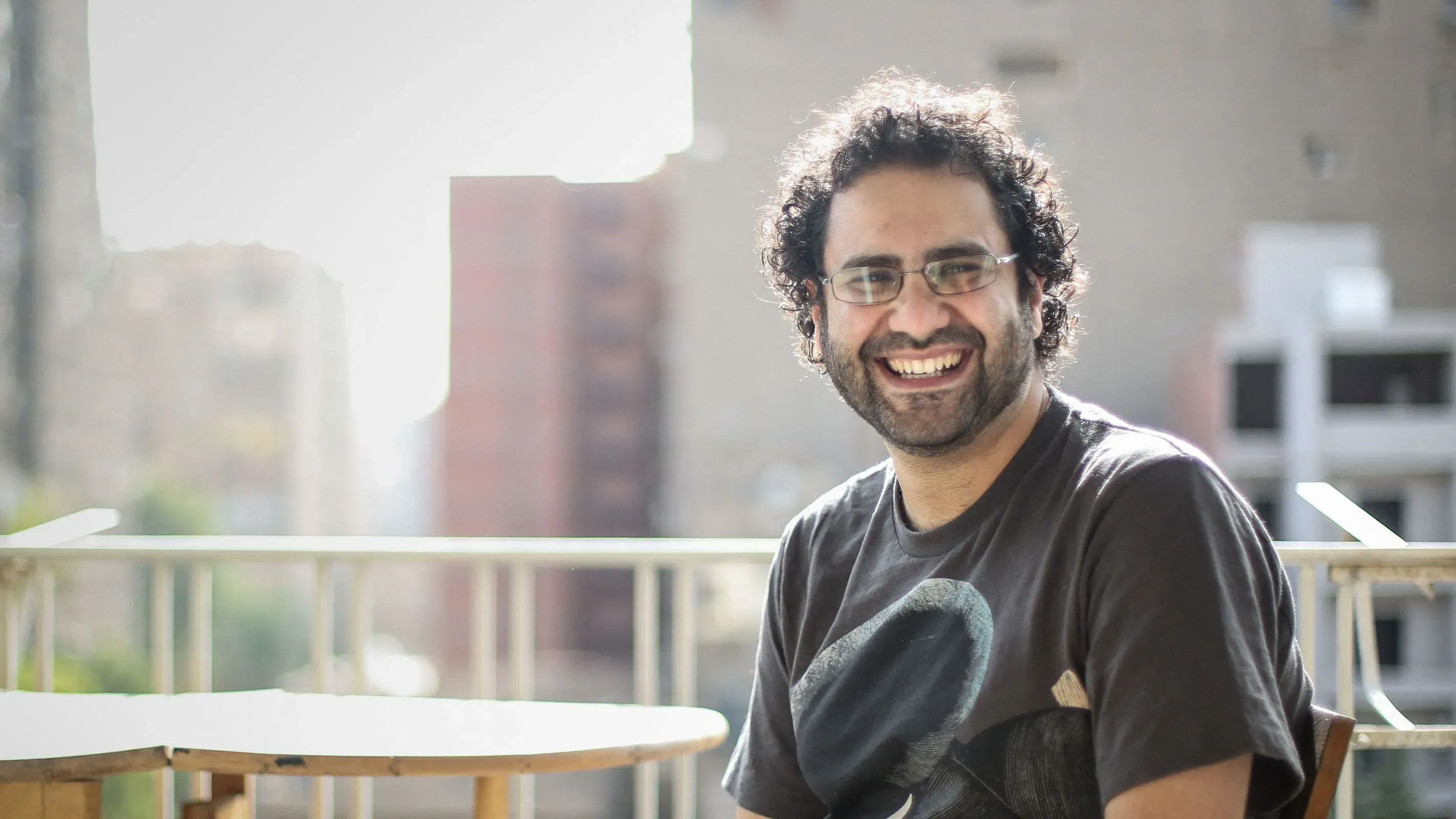 علاء عبد الفتاح، المعارض المصري المسجون، مبتسم على شرفة، وسط خلفية حضرية، يعكس حالة من الأمل رغم تدهور صحته.