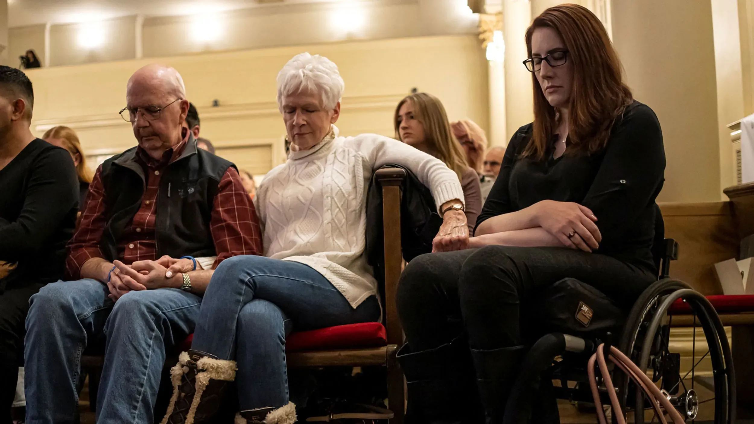 مجموعة من الأشخاص يجلسون في قاعة، مع التركيز على امرأة في كرسي متحرك، يتبادلون الأيدي في لحظة تأمل.