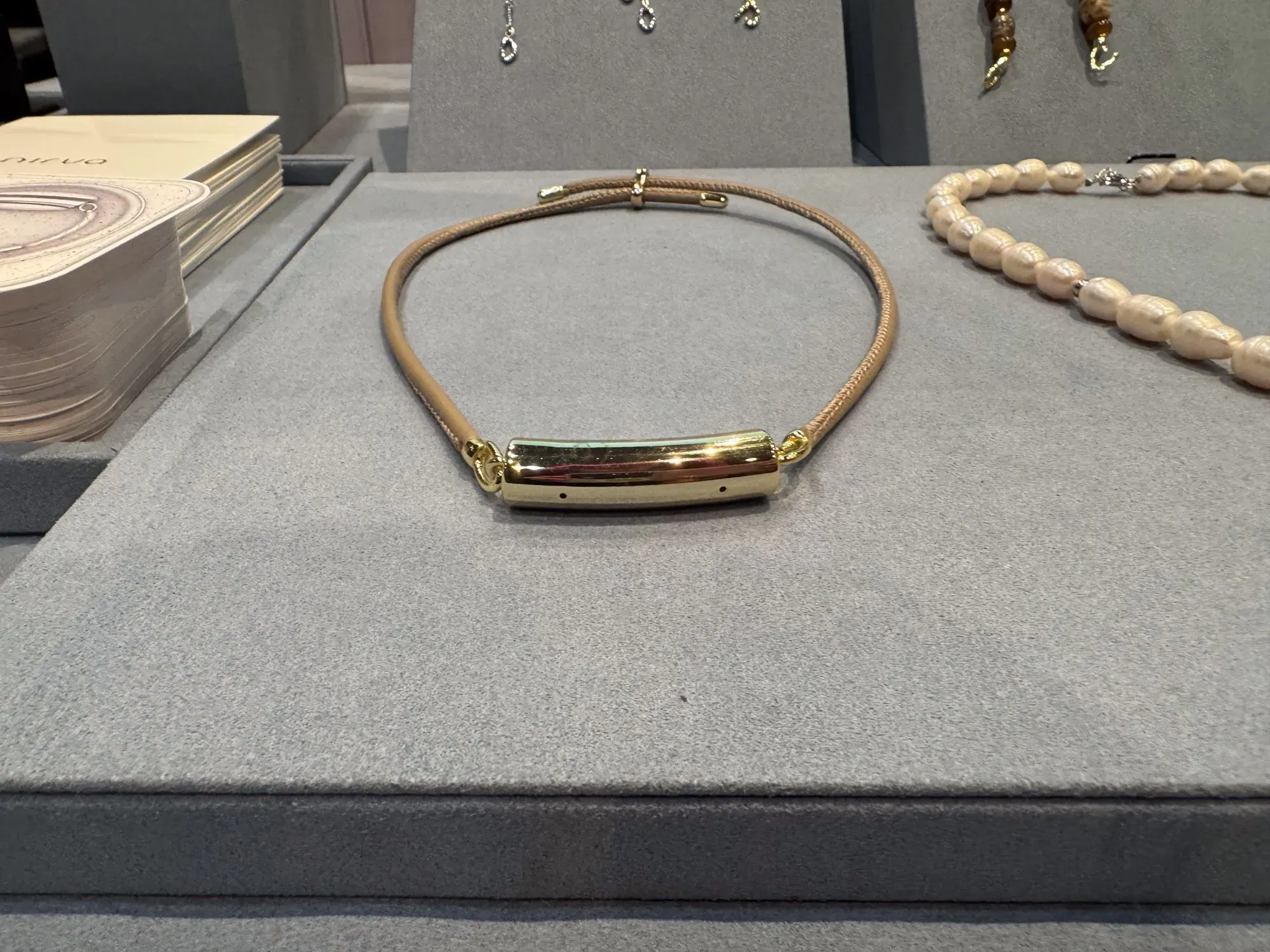 قلادة أنيقة مصنوعة من الجلد والذهب، مع تصميم بسيط، موضوعة على سطح رمادي، بجانب مجوهرات أخرى في معرض الإلكترونيات الاستهلاكية.