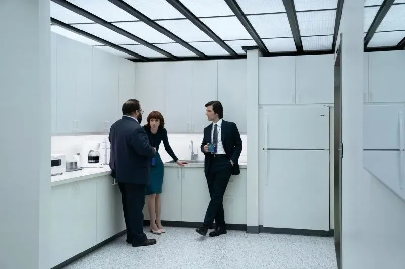 مشهد من مسلسل "Severance" يظهر ثلاثة شخصيات في مطبخ عمل حديث، يتبادلون الحديث حول تحديات العمل وثقافة الشركات.