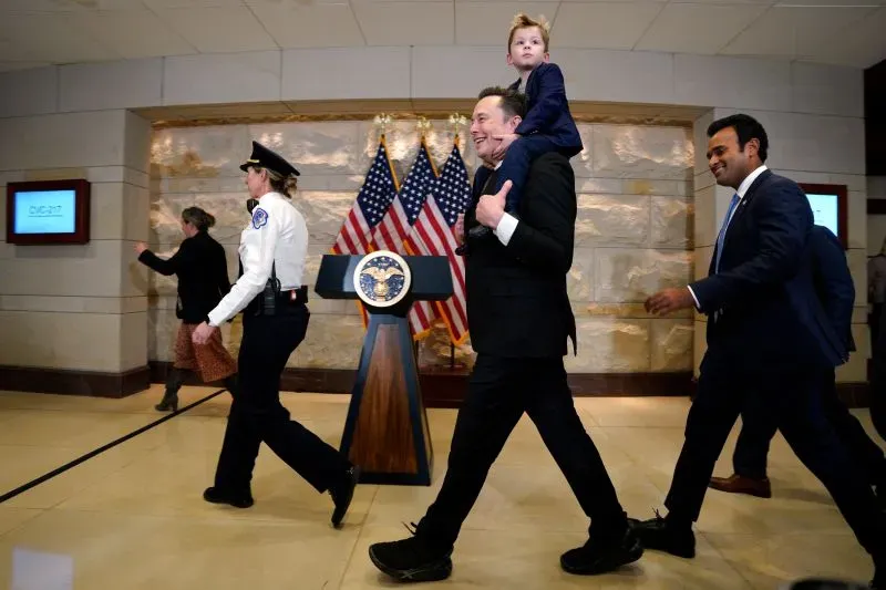 إيلون ماسك يحمل طفلاً على كتفه، بينما يسير مع فريقه في قاعة رسمية، خلفهم أعلام أمريكية ومنصة للإعلانات.