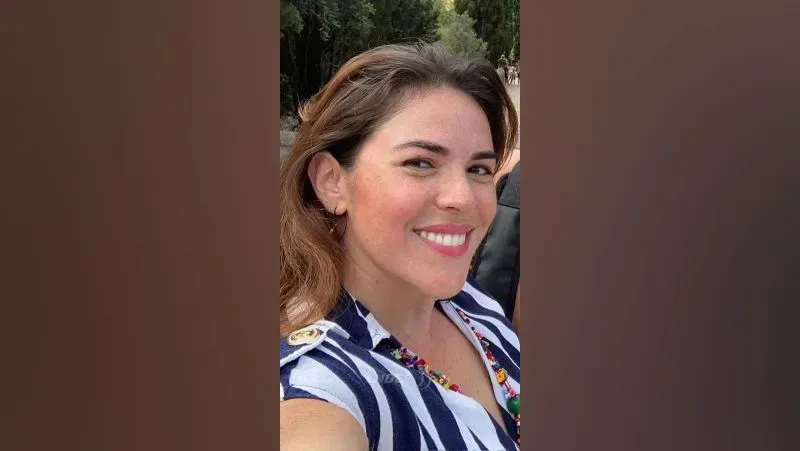 امرأة مبتسمة ترتدي قميصًا مخططًا، تلتقط صورة سيلفي في مكان خارجي، تعكس لحظة من السعادة في سياق قضية اختفاء زوجها.