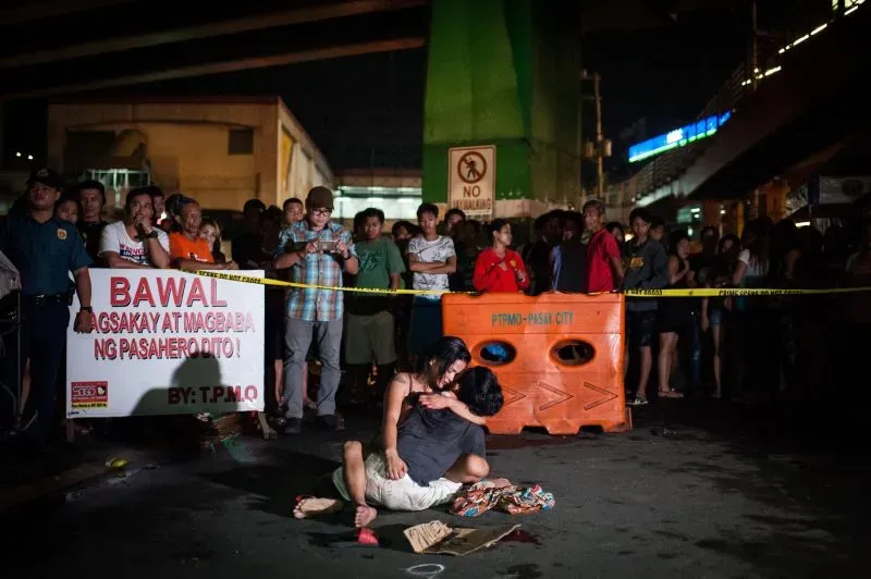 مشهد مؤثر لأسرة تتجمع حول موقع جريمة قتل، حيث يظهر شخصان يجلسان على الأرض في حالة من الحزن، بينما يتواجد جمهور من الناس خلف حواجز الشرطة.