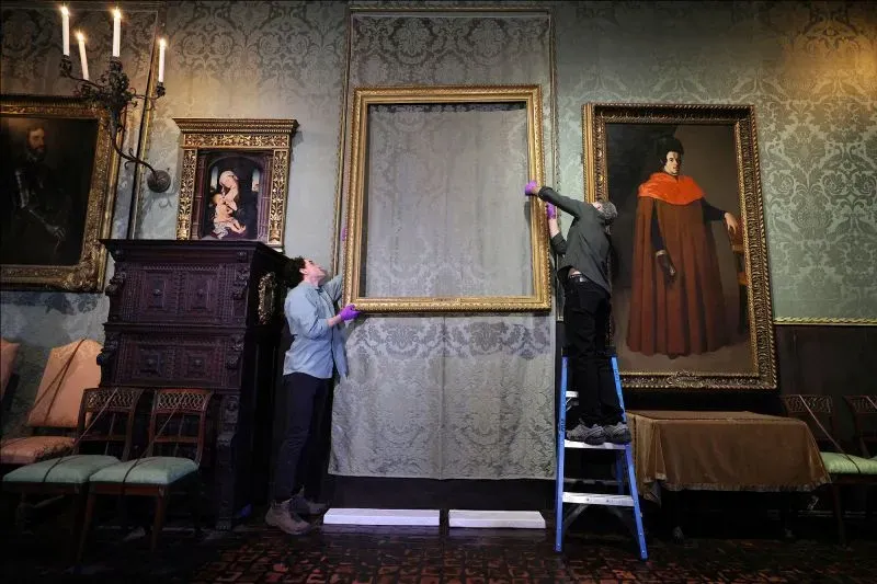 عمال يقومون بإزالة ستارة من إطار فارغ في معرض فني، بينما تظهر لوحات شهيرة على الحائط، مما يعكس تحديات سرقة الفن.