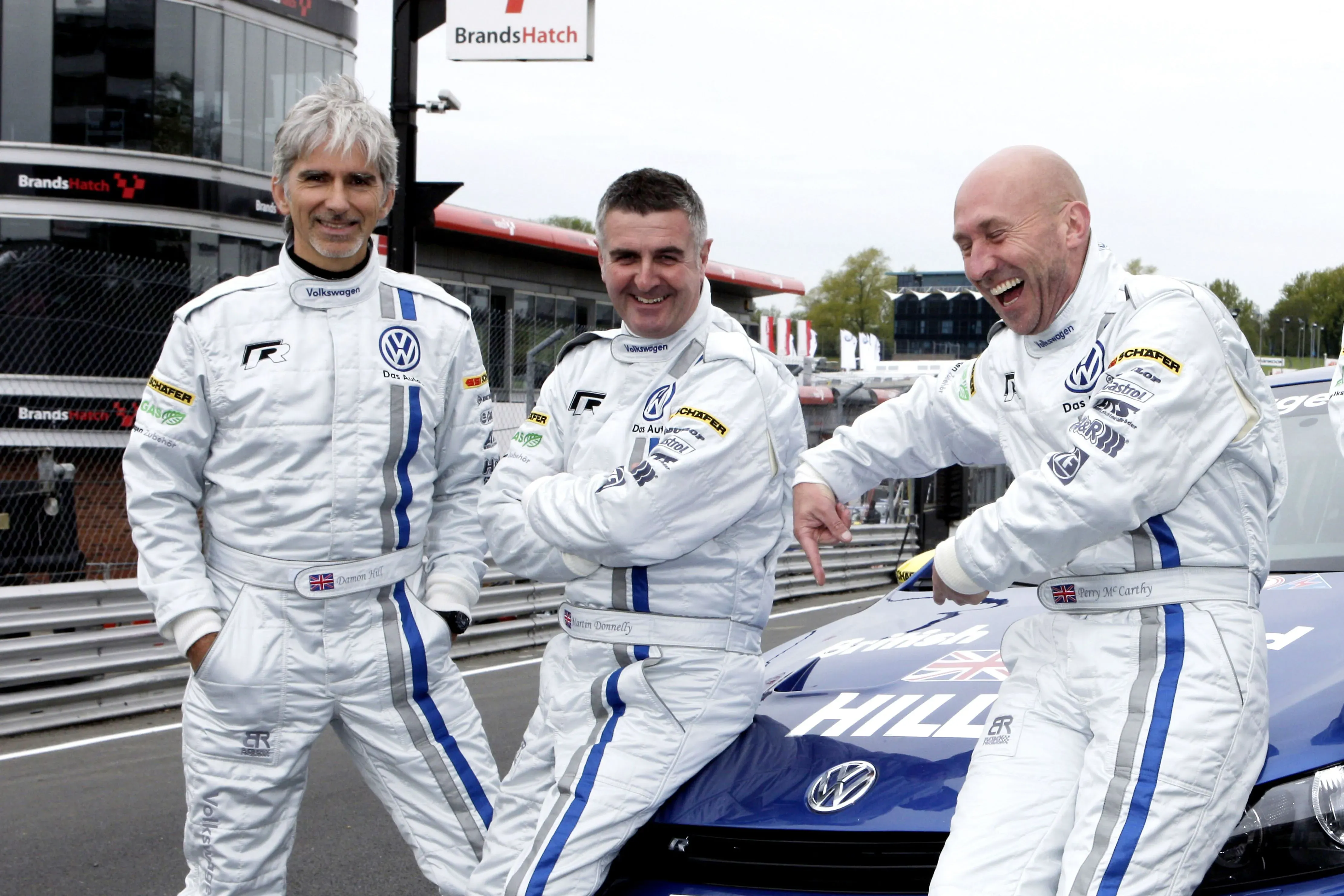 ثلاثة رجال يرتدون بدلات سباق بيضاء مع شعارات فولكس فاجن، يضحكون ويتبادلون الحديث في حلبة براندز هاتش.