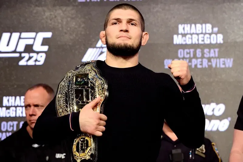 حبيب نورماغوميدوف، بطل UFC السابق، يحمل حزام البطولة ويظهر في مؤتمر صحفي، مع تعبير جاد، في خلفية تحمل شعار UFC.