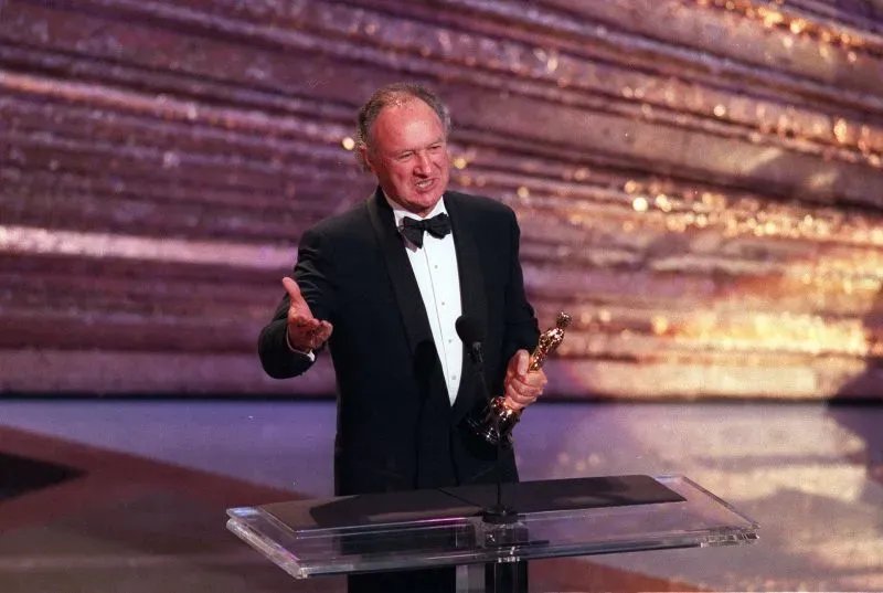 جين هاكمان يتسلم جائزة الأوسكار على المسرح، يرتدي بدلة رسمية ويعبر عن شكره، مع خلفية لامعة تعكس أجواء الحفل.