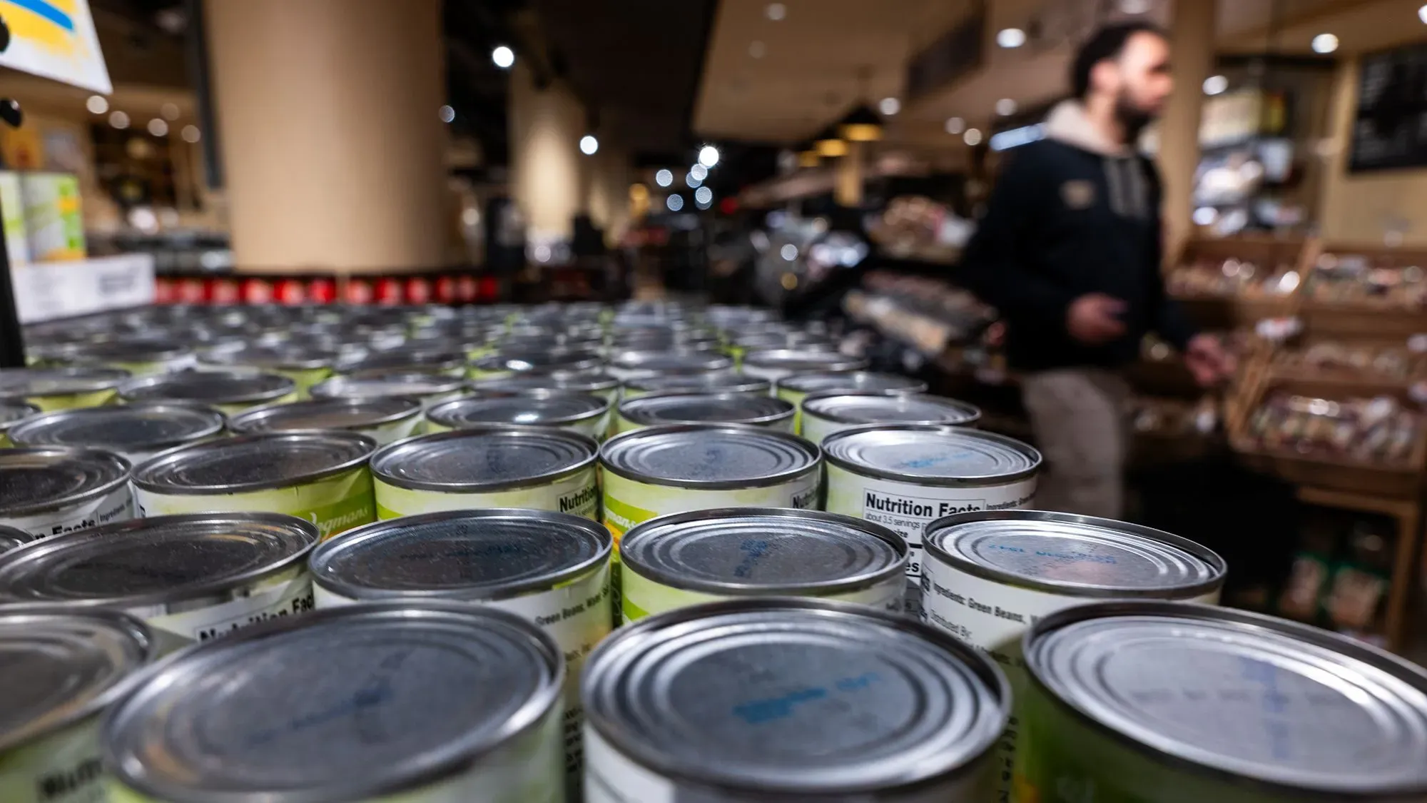 تظهر الصورة صفوفًا من علب الطعام في متجر، مما يعكس تأثير التعريفات الجمركية على أسعار المنتجات الاستهلاكية.