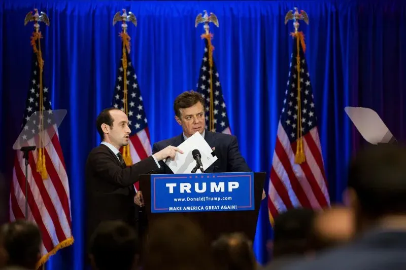 ستيفن ميلر، مستشار ترامب، يتحدث مع مساعده أثناء مؤتمر صحفي، مع خلفية من الأعلام الأمريكية ولافتة "ترامب".