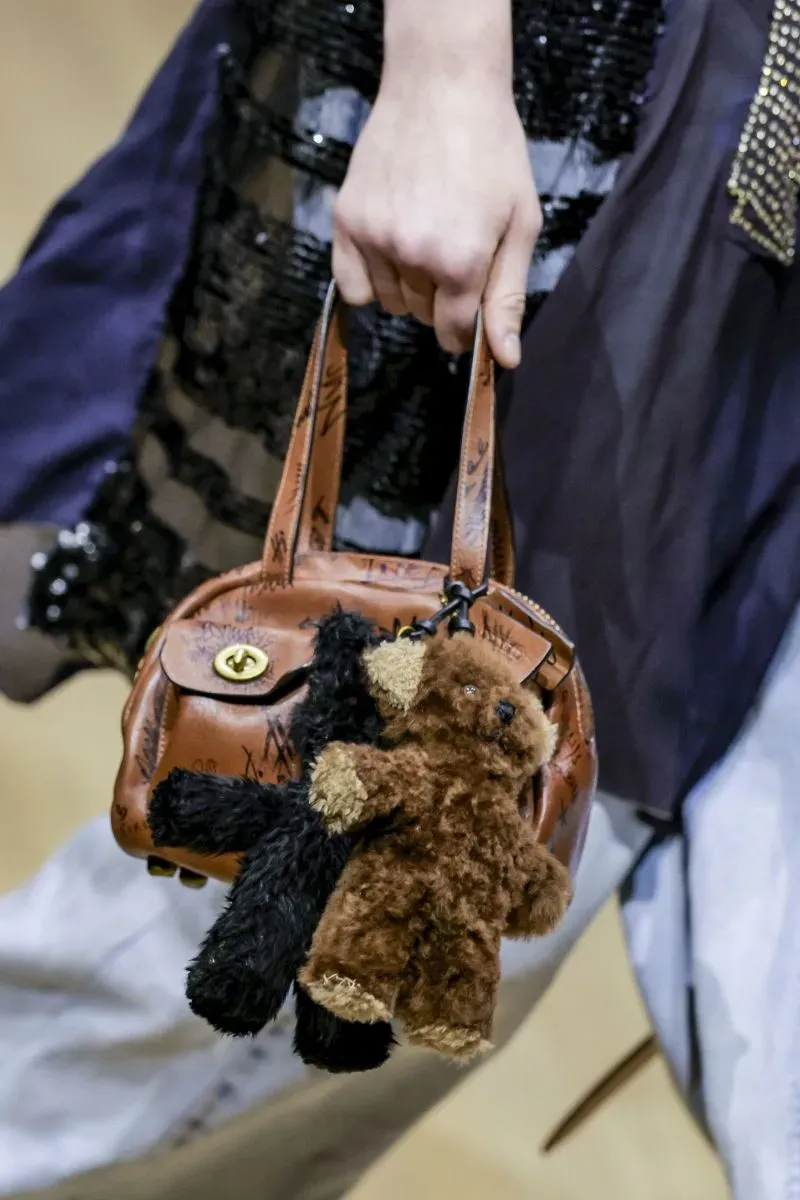 حقيبة يد جلدية بنية تحمل دميتين محشوتين صغيرتين، واحدة بنية والأخرى سوداء، تعكس اتجاهات الموضة المعاصرة.