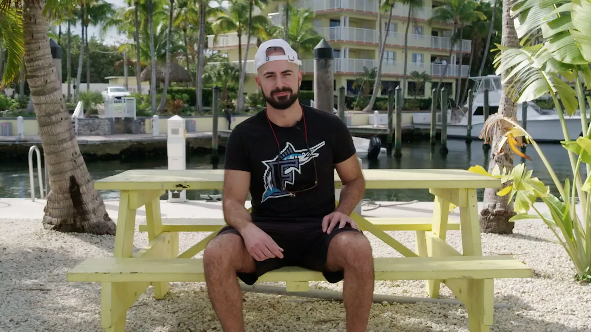 رجل يجلس على طاولة صفراء في منطقة خارجية بجوار المياه في فلوريدا كيز، مع منازل وقوارب في الخلفية.