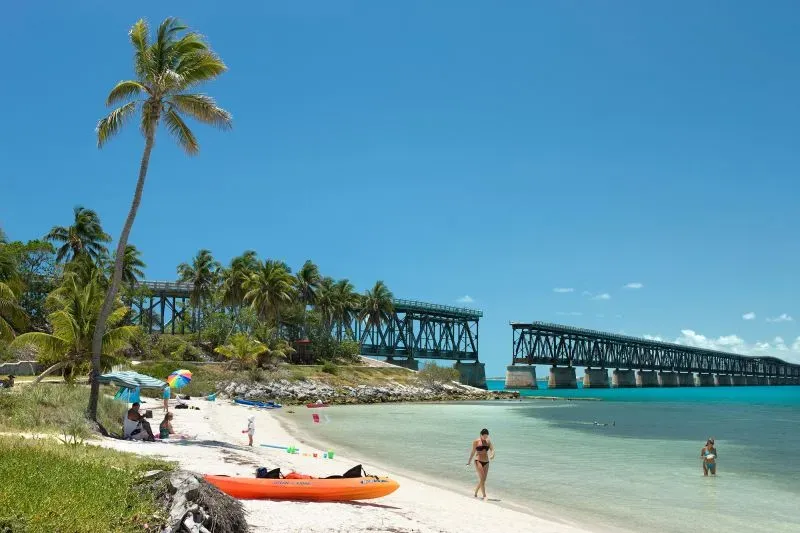 شاطئ في فلوريدا كيز مع أشجار النخيل، وقوس من الجسر الحديدي في الخلفية، وزوار يستمتعون بالماء والشاطئ.
