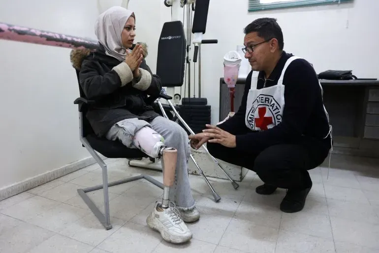 شابة ترتدي طرفًا صناعيًا تتحدث مع موظف من الصليب الأحمر في مركز طبي، تعبيرًا عن الحاجة الملحة للرعاية الصحية في غزة.