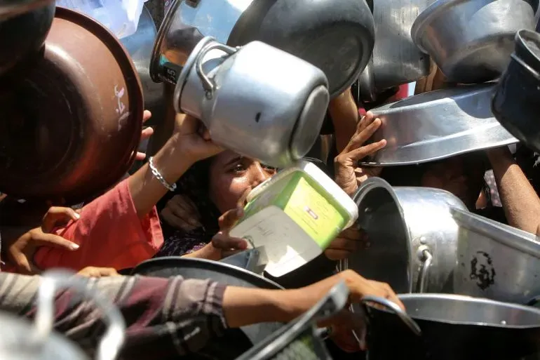 تظهر الصورة مجموعة من الأشخاص يتجمعون حول حاويات الطعام، مما يعكس معاناتهم من نقص الغذاء في ظل الأزمات الإنسانية المتزايدة.