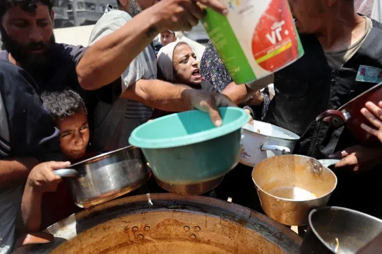 تجمع حشد من الناس في غزة حول وعاء كبير للحصول على الطعام، مع تعبيرات يأس وحاجة واضحة، مما يعكس أزمة الغذاء المتفاقمة في المنطقة.
