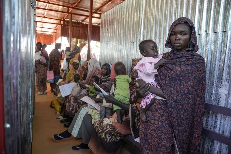 نساء وأطفال يجلسون في انتظار المساعدة في مركز إغاثة، يعكس معاناة المدنيين في السودان بسبب الصراع المستمر.