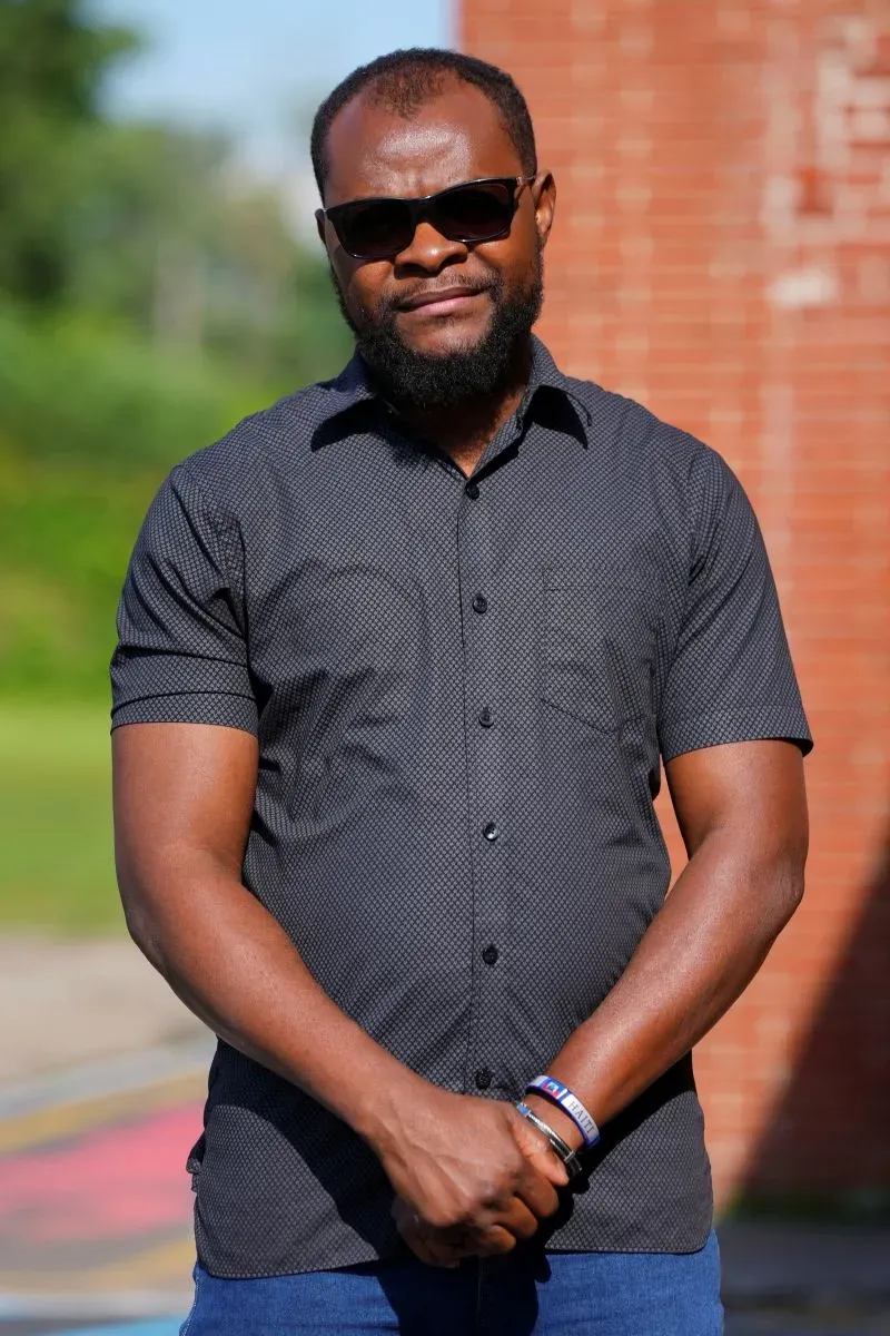 رجل يرتدي نظارات شمسية وقميصًا أسود، يقف في مكان مفتوح مع خلفية غير واضحة، معبرًا عن قلقه بشأن وضع المهاجرين الهايتيين في الولايات المتحدة.