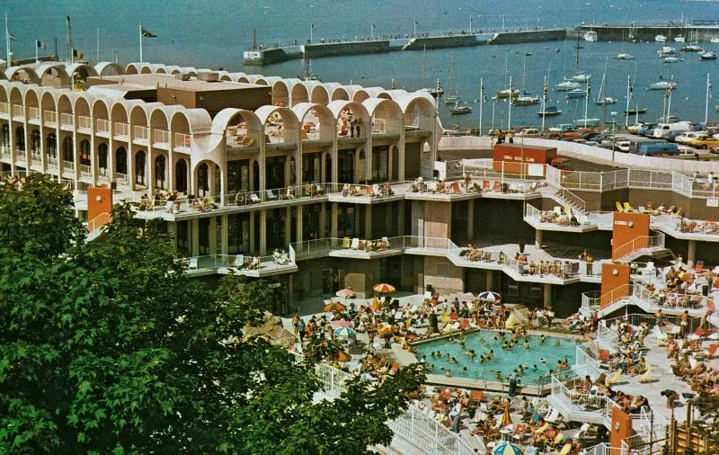 مجموعة من الناس يستمتعون بأشعة الشمس حول مسبح في منتجع ساحلي بريطاني، مع مناظر للبحر والقوارب في الخلفية.