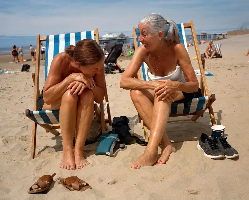 امرأتان تجلسان على الكرسي الشاطئي، تتبادلان الحديث والضحك على شاطئ بحر، مع مشهد البحر في الخلفية.