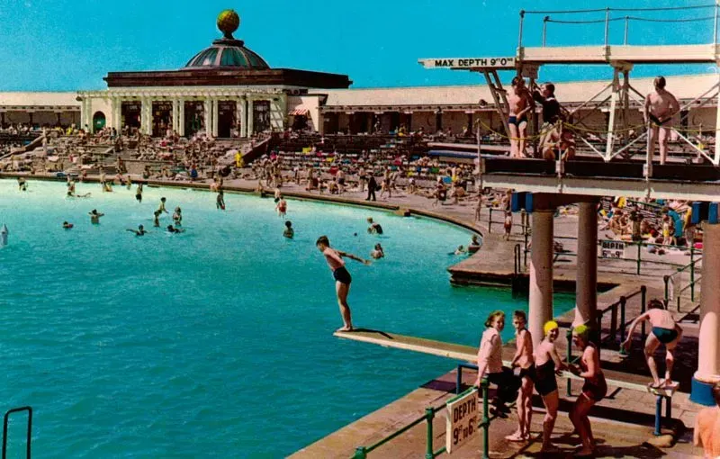 مجموعة من الأشخاص يستمتعون بالسباحة في مسبح كبير على شاطئ البحر، مع وجود منصة للغطس خلفهم، تعكس أجواء الصيف البريطاني.