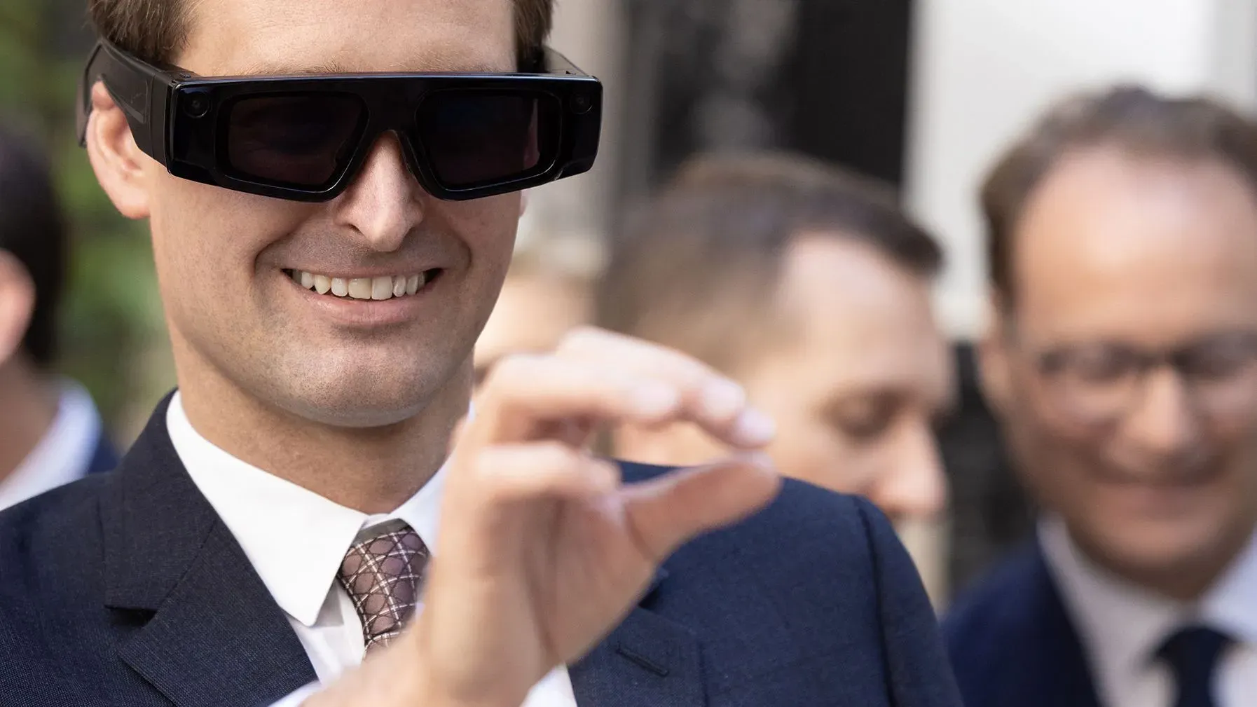 رجل يرتدي نظارات ذكية جديدة، مبتسمًا ويظهر إيماءة بيده، مع مجموعة من الأشخاص في الخلفية. تعكس النظارات التطورات في تقنية الذكاء الاصطناعي.