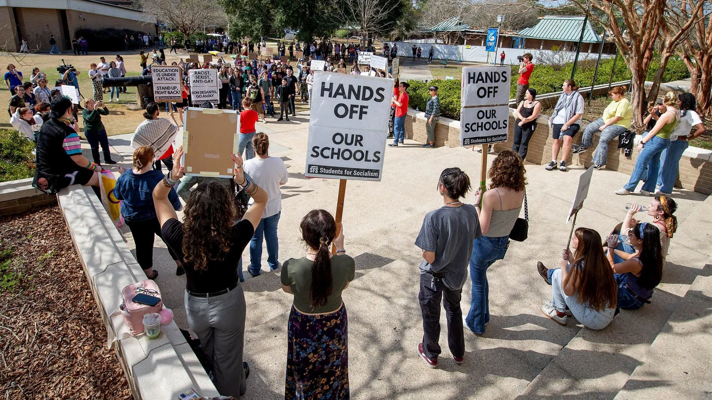 محتجون يحملون لافتات تعبر عن رفضهم لتخفيضات الإنفاق في التعليم، وسط تجمع في حرم جامعي، مما يعكس القلق حول مستقبل التعليم.