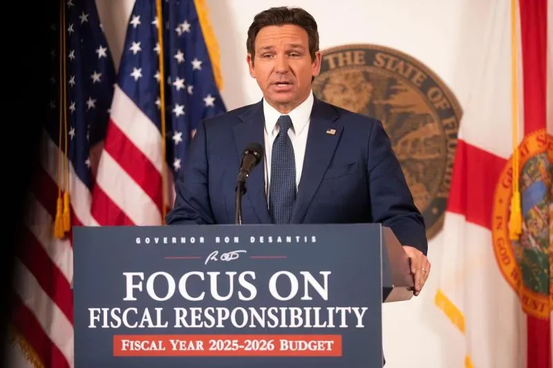 حاكم فلوريدا رون ديسانتس يتحدث في مؤتمر صحفي حول الميزانية، مع التركيز على المسؤولية المالية، خلفه أعلام الولاية.