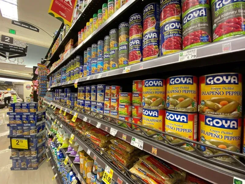 رفوف متجر تحتوي على علب طعام متنوعة من علامة جويا، بما في ذلك الفاصوليا والفلفل الحار، تعكس شهرة منتجات الشركة.