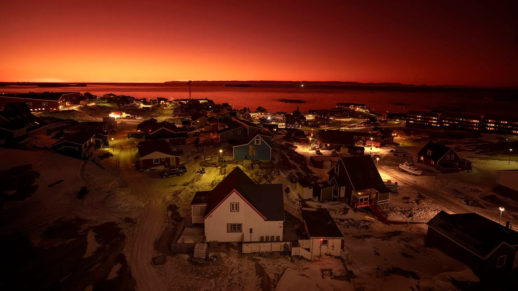 منظر ليلي لبلدة في غرينلاند، تظهر المنازل الملونة مضاءة تحت سماء غائمة باللون البرتقالي، مما يعكس جمال الطبيعة القطبية.