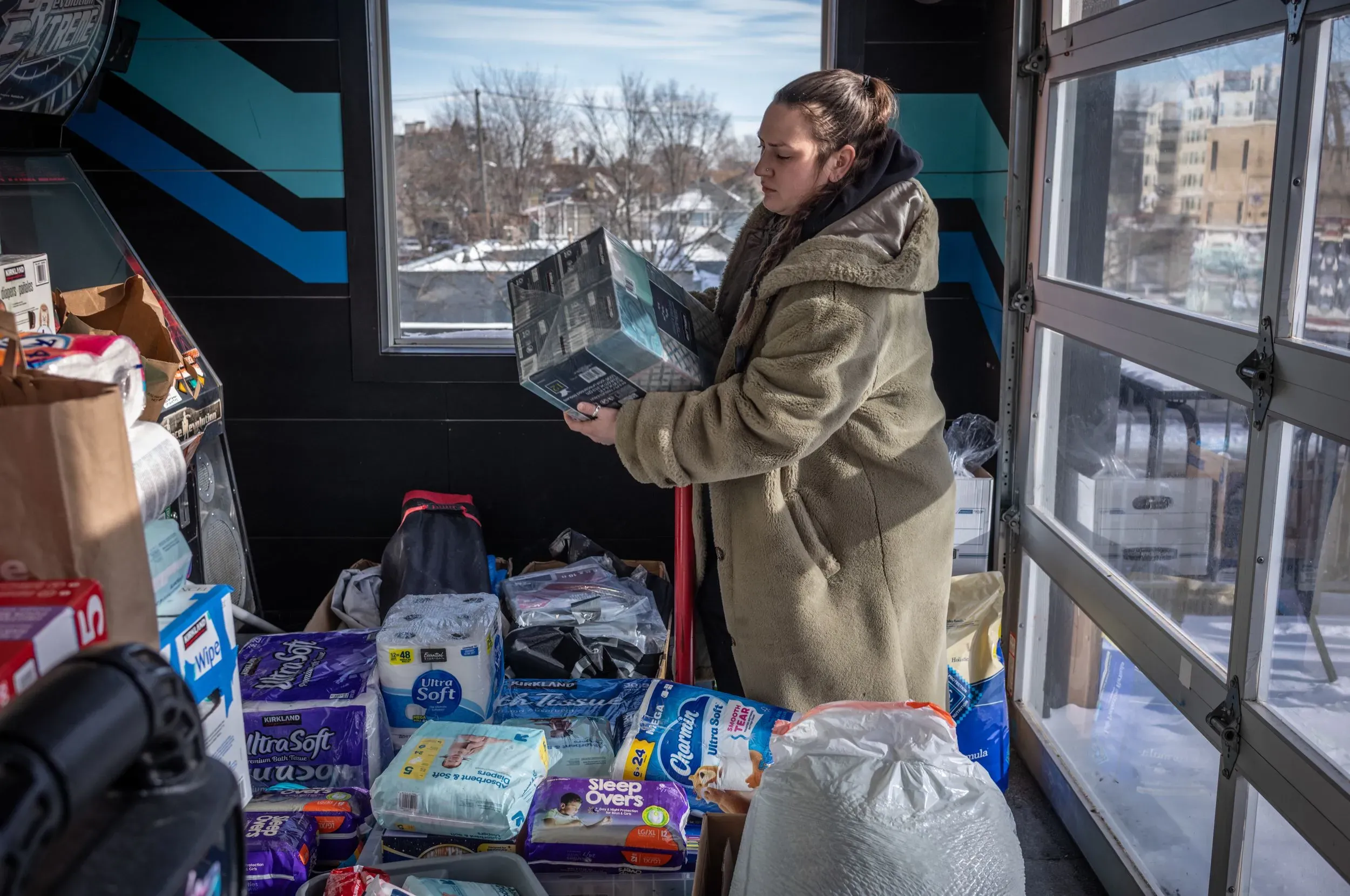 امرأة ترتدي معطفًا سميكًا، تقوم بترتيب صناديق من الإمدادات، مثل الحفاضات والمناديل، في مكان مخصص لتوزيع المساعدات في الشتاء.
