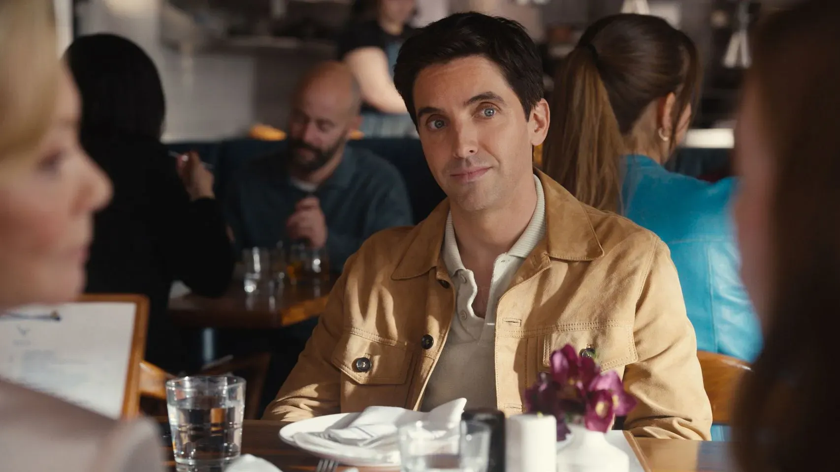 لقطة من مسلسل "Hacks" تظهر شخصية جيمي لوساكي وهو جالس في مطعم، مع زهور على الطاولة، يعكس لحظة من التفاعل الكوميدي.