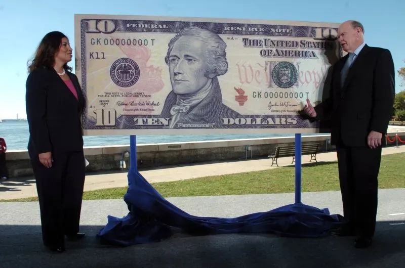 سيدة ورجل يقفان بجانب لافتة كبيرة لورقة نقدية من فئة 10 دولارات، مع صورة ألكسندر هاملتون، في حدث يركز على تغييرات العملة.