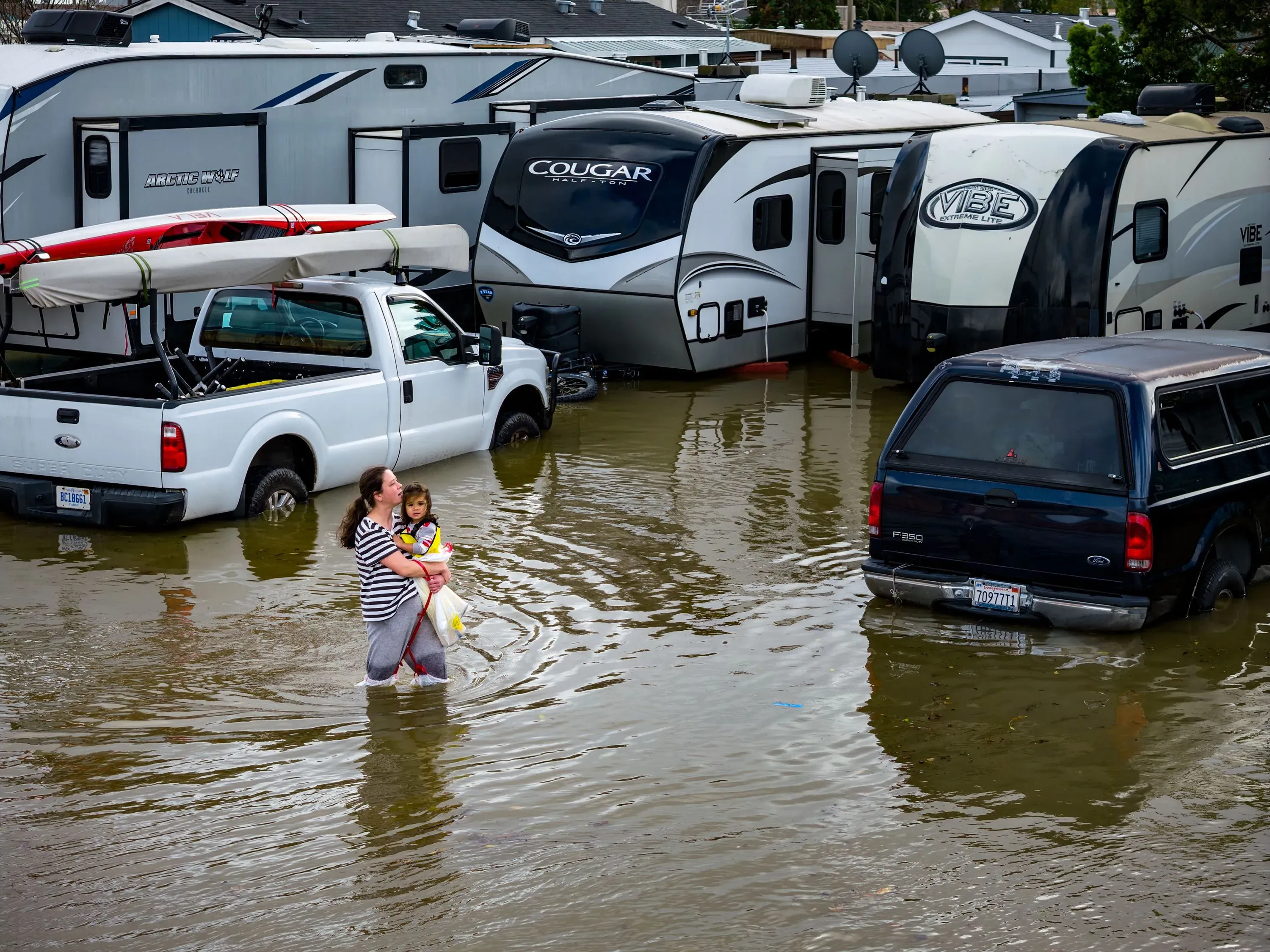 امرأة تحمل طفلاً في مياه الفيضانات، محاطة بمركبات غارقة، تعكس تأثير الأمطار الغزيرة والمد والجزر الملكي في شمال كاليفورنيا.