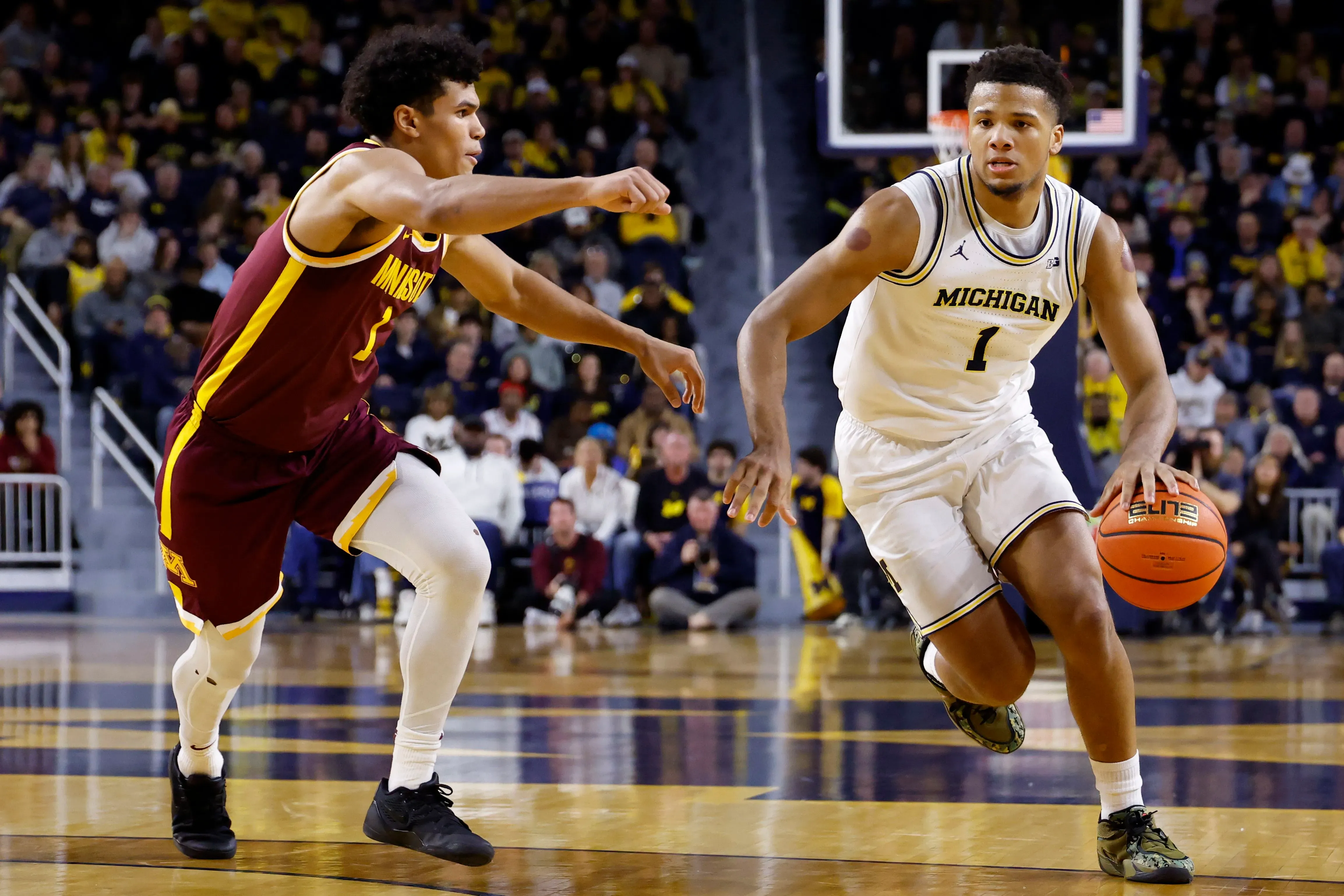 لاعب كرة سلة من فريق ميشيغان يحمل الكرة ويتجه نحو السلة، بينما يواجه مدافعًا من فريق ميني سوتا في مباراة تنافسية.