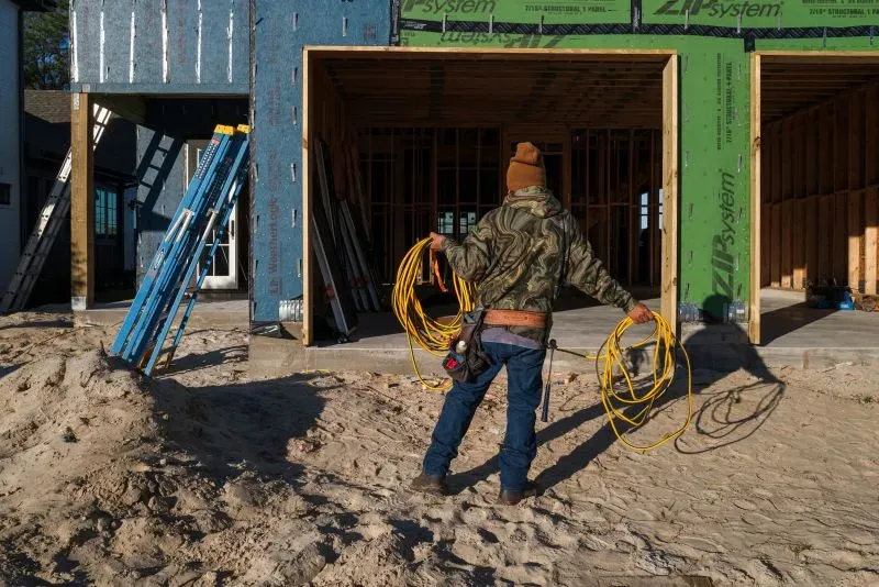 عامل بناء يحمل أسلاك كهربائية في موقع بناء منزل جديد، مع وجود سقالات وأجزاء من الهيكل الظاهر خلفه.