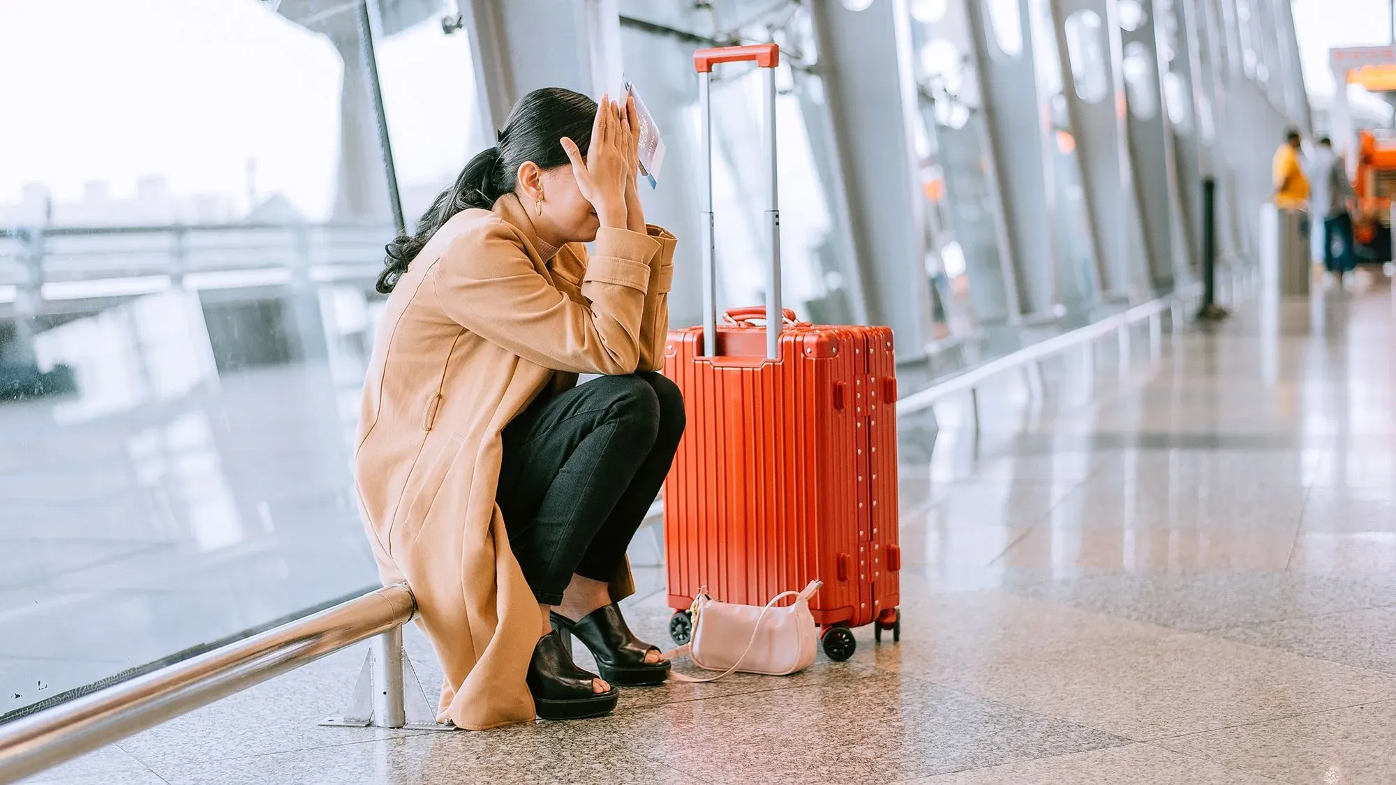 امرأة تجلس في مطار، تعبر عن الإحباط والقلق، بجانب حقيبة سفر حمراء. تعكس الصورة مشاعر التوتر المرتبطة بالسفر بالطائرة.