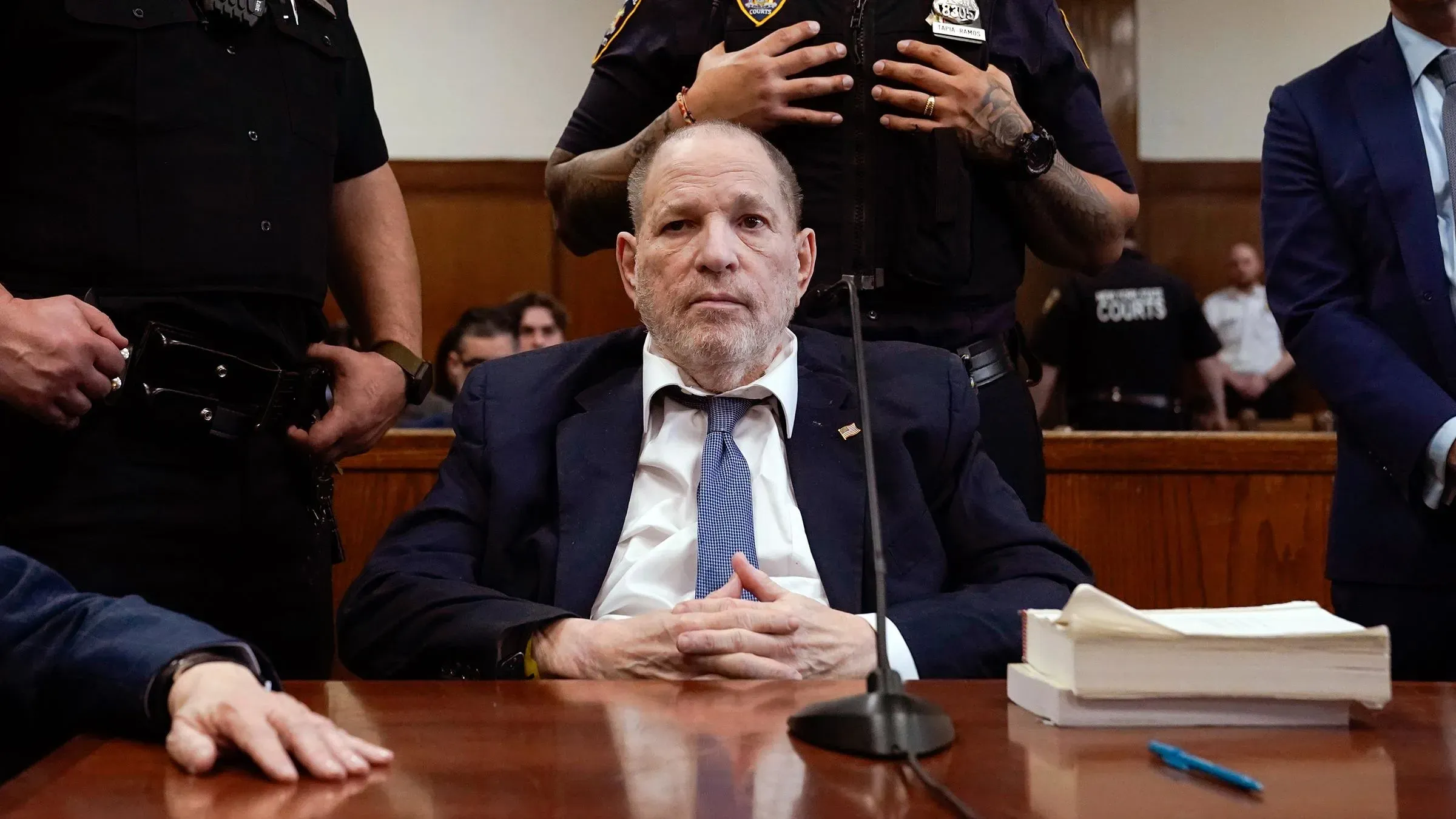 هارفي واينستين جالس في قاعة المحكمة محاطًا بالشرطة، مع التركيز على تعبير وجهه الجاد، في سياق إعادة محاكمته بتهم الاعتداء الجنسي.