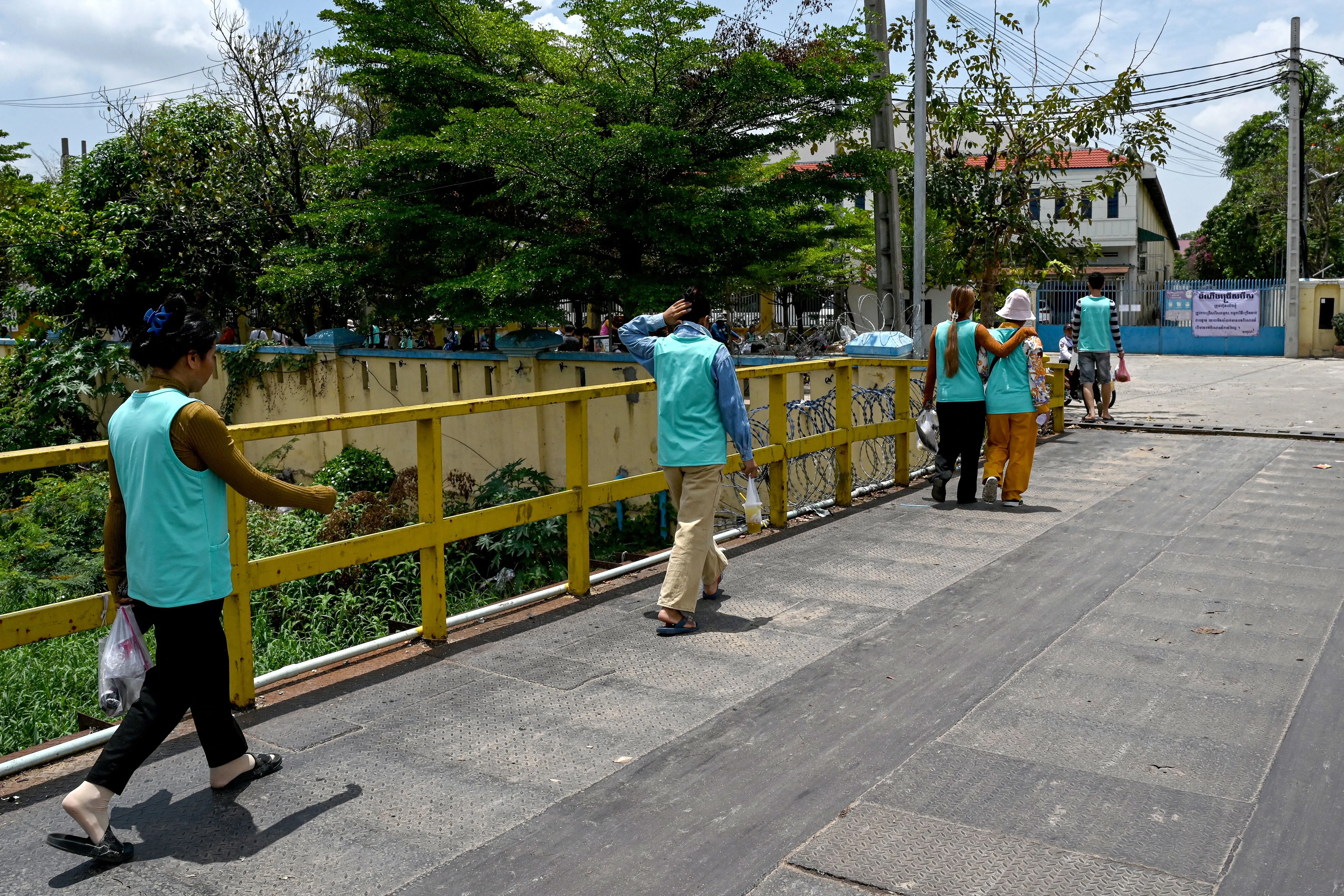 مجموعة من الأشخاص يرتدون قمصان زرقاء، يسيرون على جسر في كمبوديا، وسط أشجار ونباتات، في سياق تأثير التعريفات الجمركية على الأعمال.
