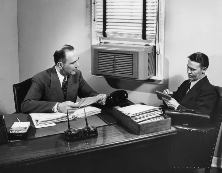 رجلان يجلسان في مكتب، مع مكيف هواء مثبت على النافذة خلفهما، يتبادلان الحديث حول الوثائق.