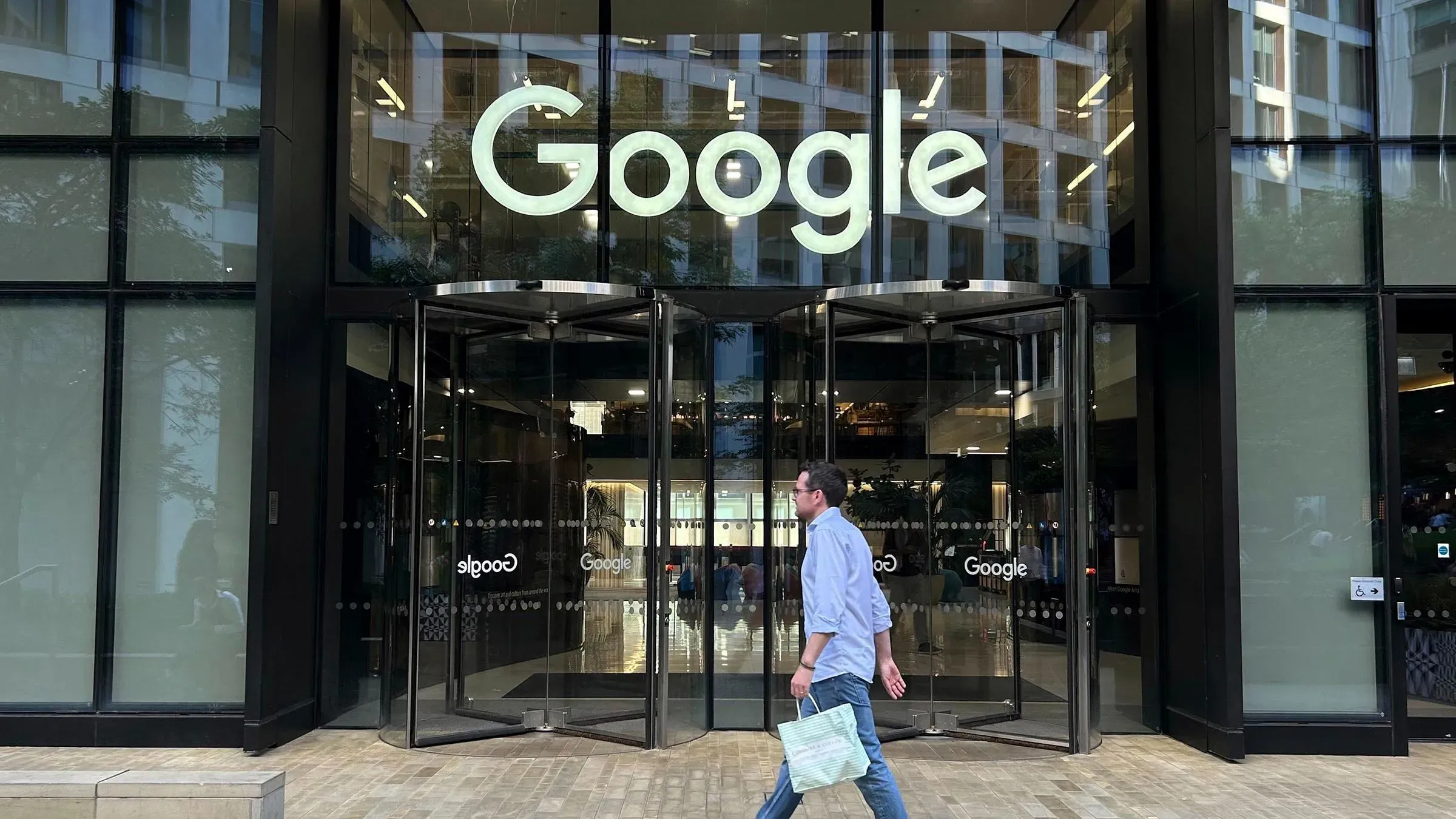 واجهة مبنى جوجل، مع شعار الشركة البارز، ورجل يسير بجوار مدخل دوار، مما يعكس هيمنة جوجل في مجال التكنولوجيا والبحث.