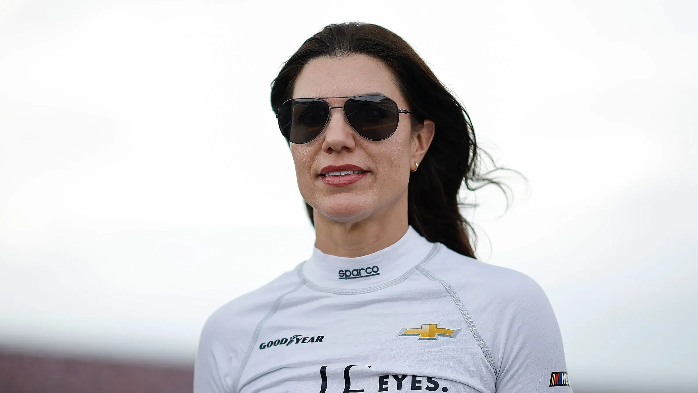 كاثرين ليج، سائقة سباقات بريطانية، ترتدي نظارات شمسية وملابس سباق، مع خلفية سماء غائمة، تعبر عن تحدياتها في عالم رياضة السيارات.